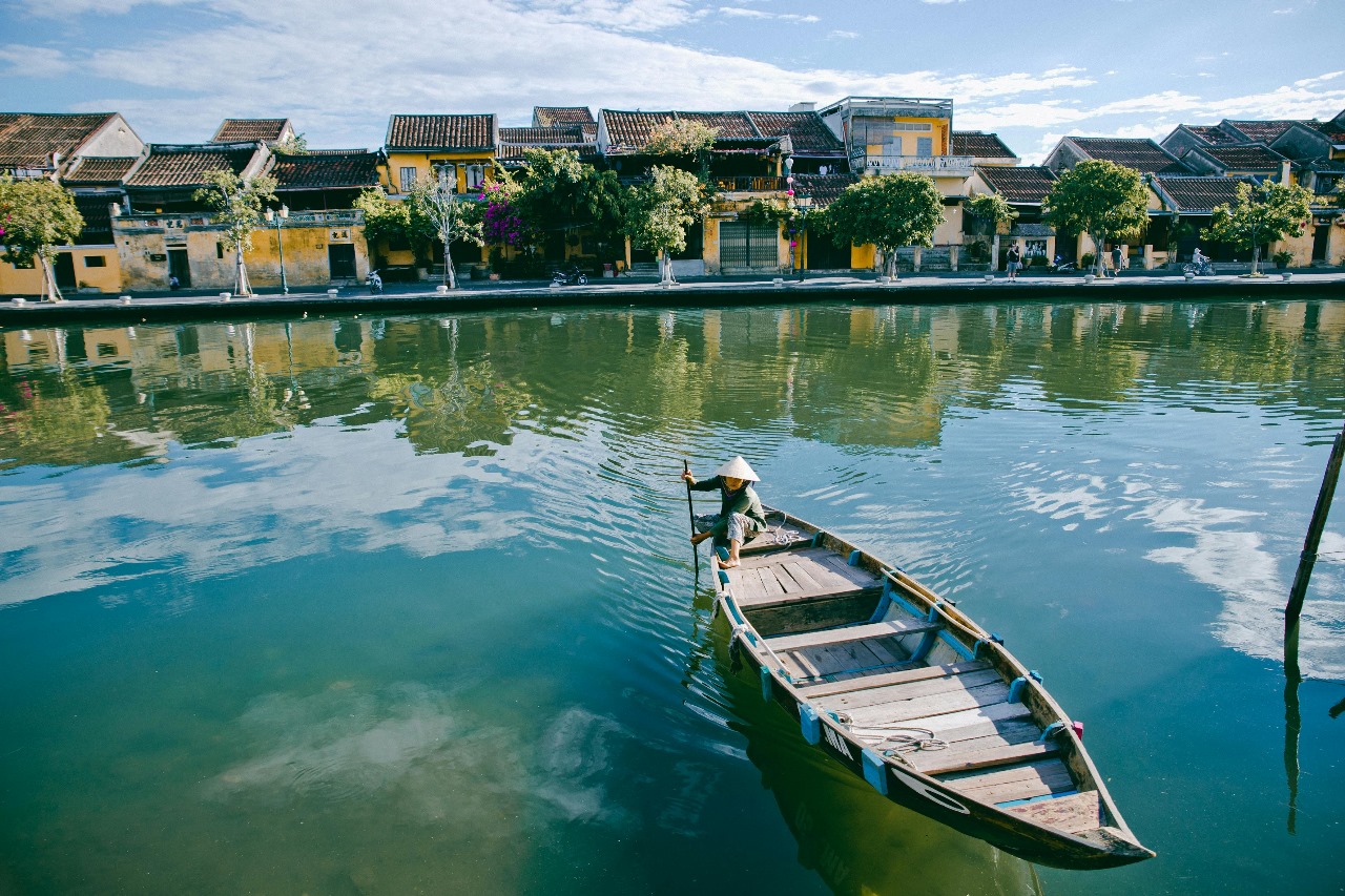 Peaceful Hoi An waterfront with traditional yellow colonial buildings reflected in calm river water and a person in conical hat rowing a wooden boat, showcasing Vietnam's cultural heritage and serene beauty